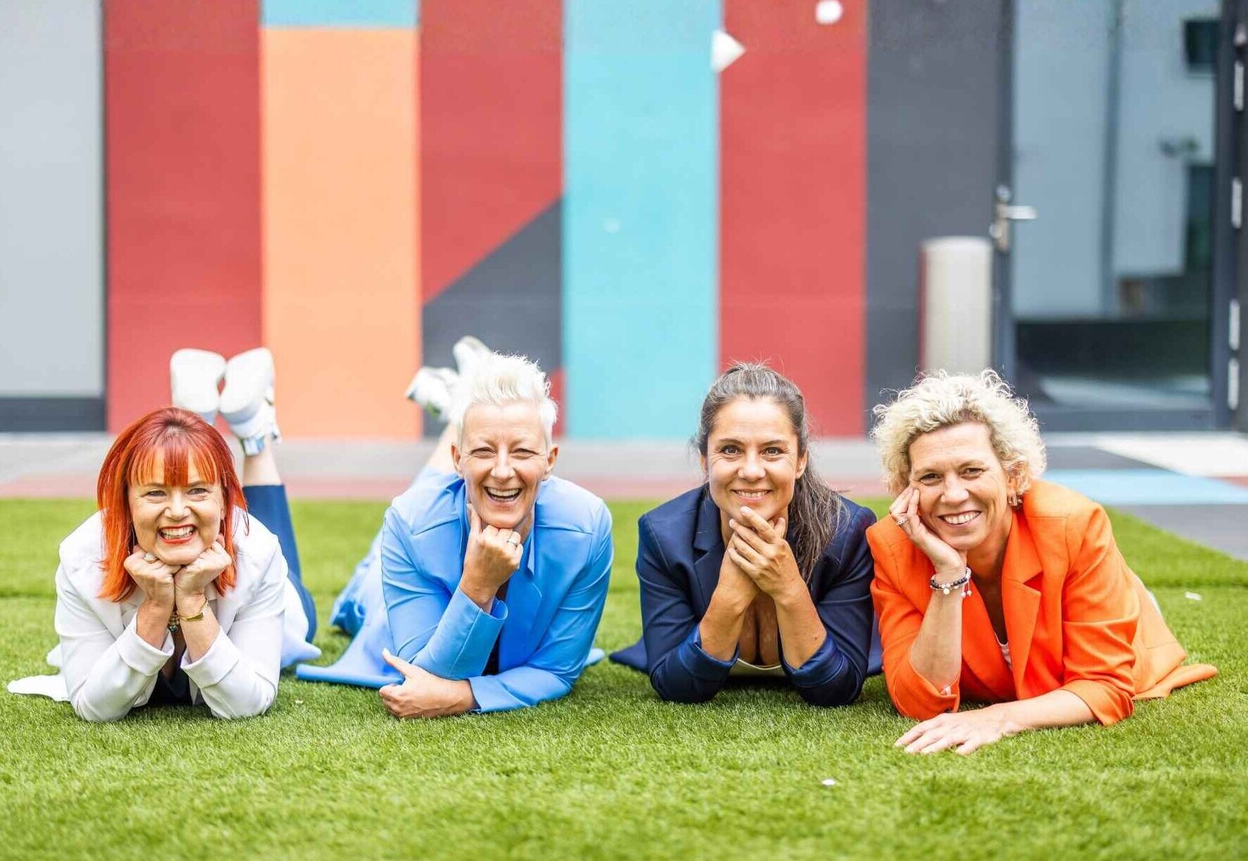 Vier Frauen liegen lachend auf einer Wiese vor einer bunten Fassade